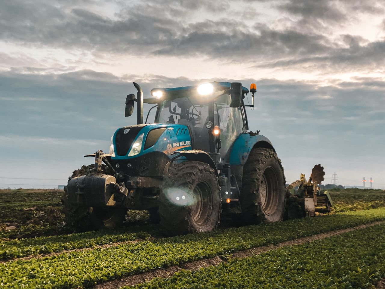 Big tractor with headlights and plow riding on green agricultural plantation in countryside on farmland against cloudy sky during harvest season